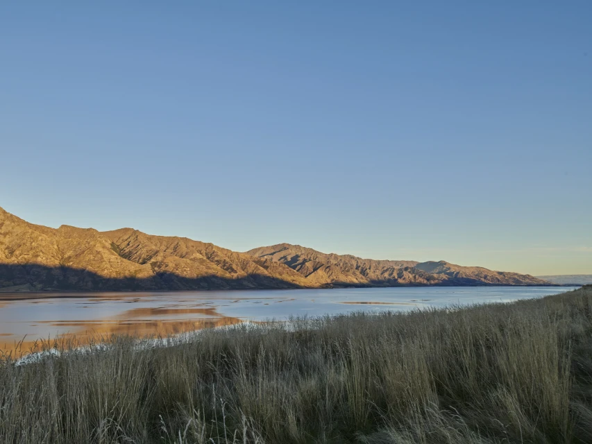 Scenic view of mountains and a lake reflecting the sky at sunset, with golden-brown grasses in the foreground, creating a tranquil landscape.