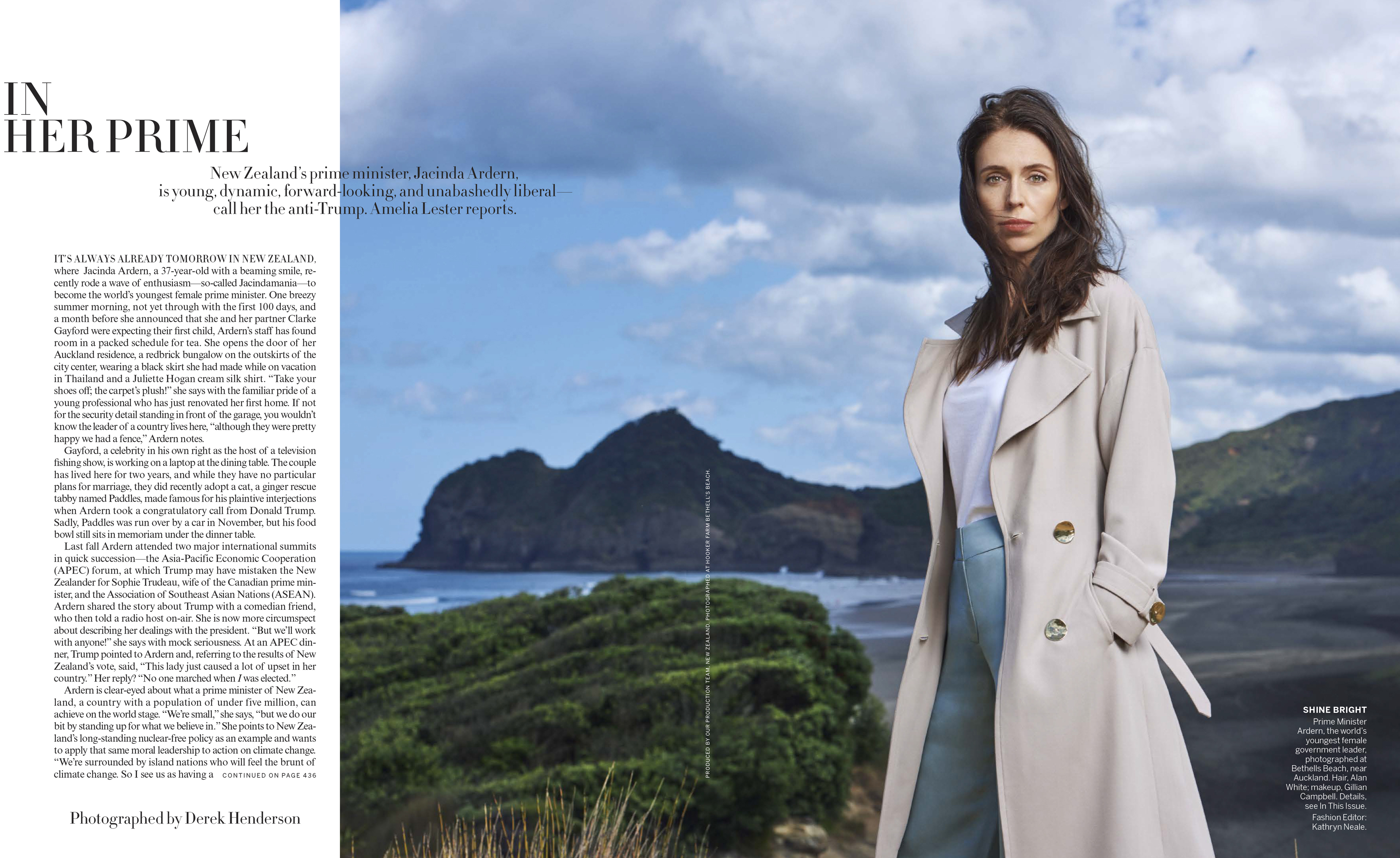 Jacinda Ardern stands in a tan coat and blue pants against a coastal backdrop. It is daytime, and the photo has soft, natural lighting.