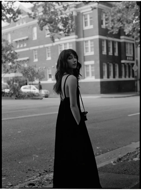 Monochrome portrait of a young woman in a black dress standing on a city street with a building and trees in the background.