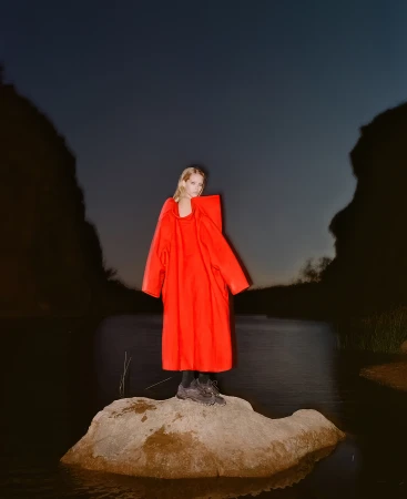 Model in a bright red coat standing on a rocky outcrop surrounded by dark water under an evening sky, photographed by Derek Henderson