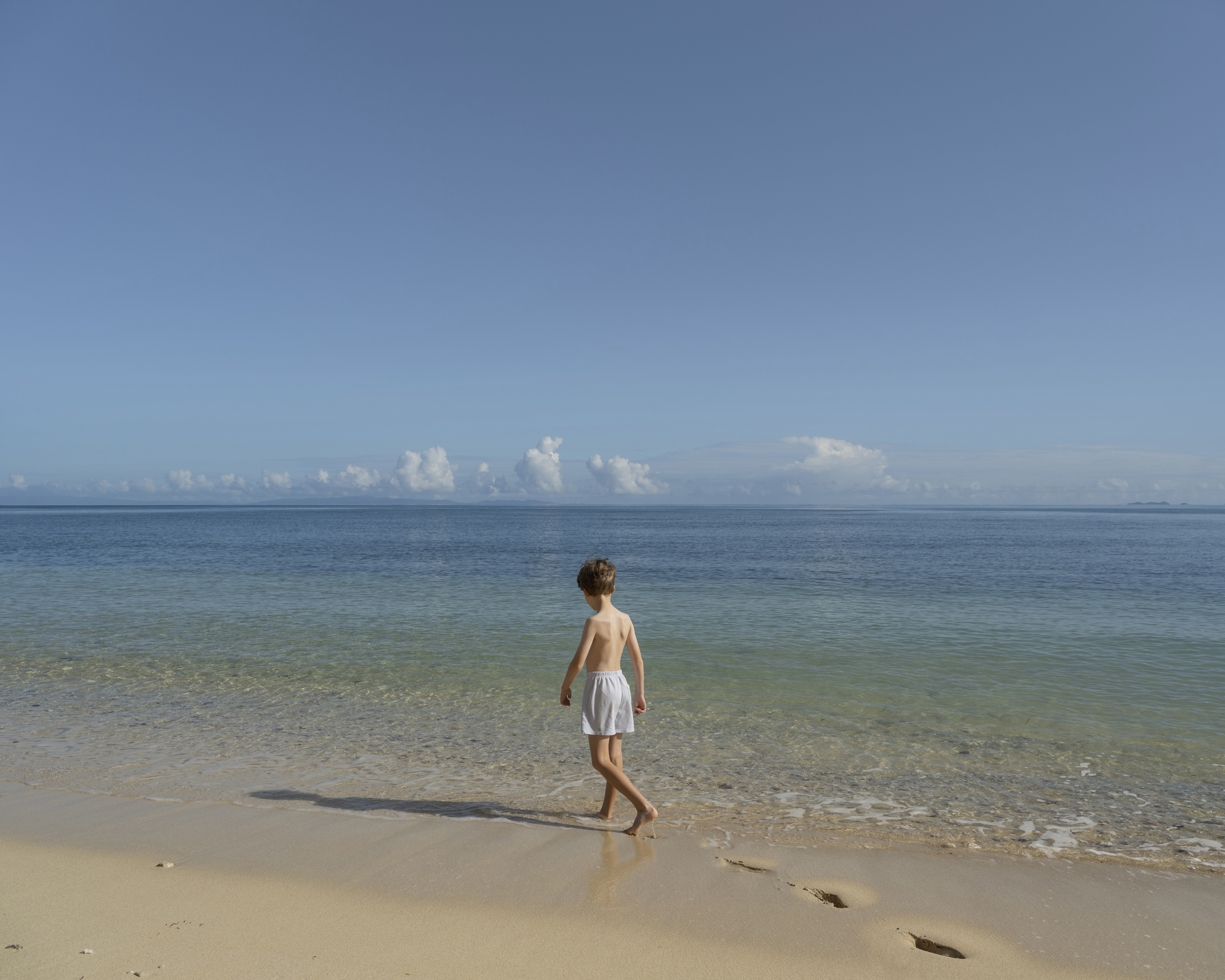 A child walks along a sandy beach with clear blue ocean water under a bright, sunny sky. Footprints mark the sand.
