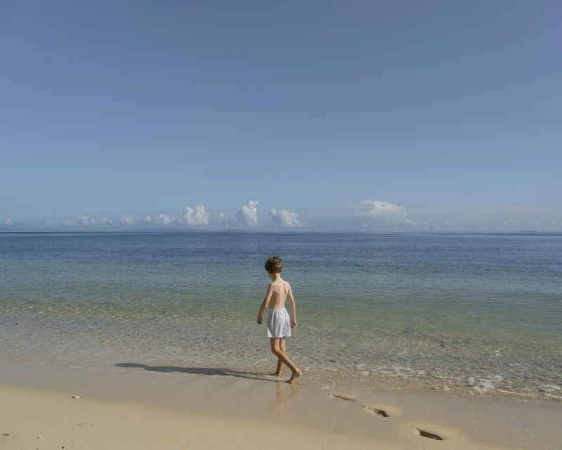 A child walks along a sandy beach with clear blue ocean water under a bright, sunny sky. Footprints mark the sand.