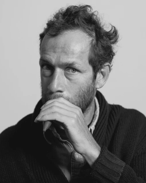 Black and white portrait of an adult male with a beard, hand near his mouth, and wearing a dark cardigan against a plain background.