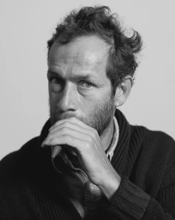 Black and white portrait of an adult male with a beard, hand near his mouth, and wearing a dark cardigan against a plain background.