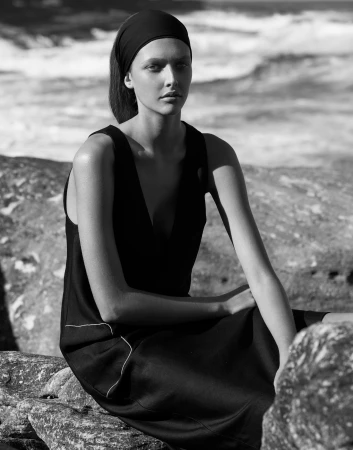 Black and white portrait of a woman in a black dress and headband seated outdoors on rocky terrain near the ocean.