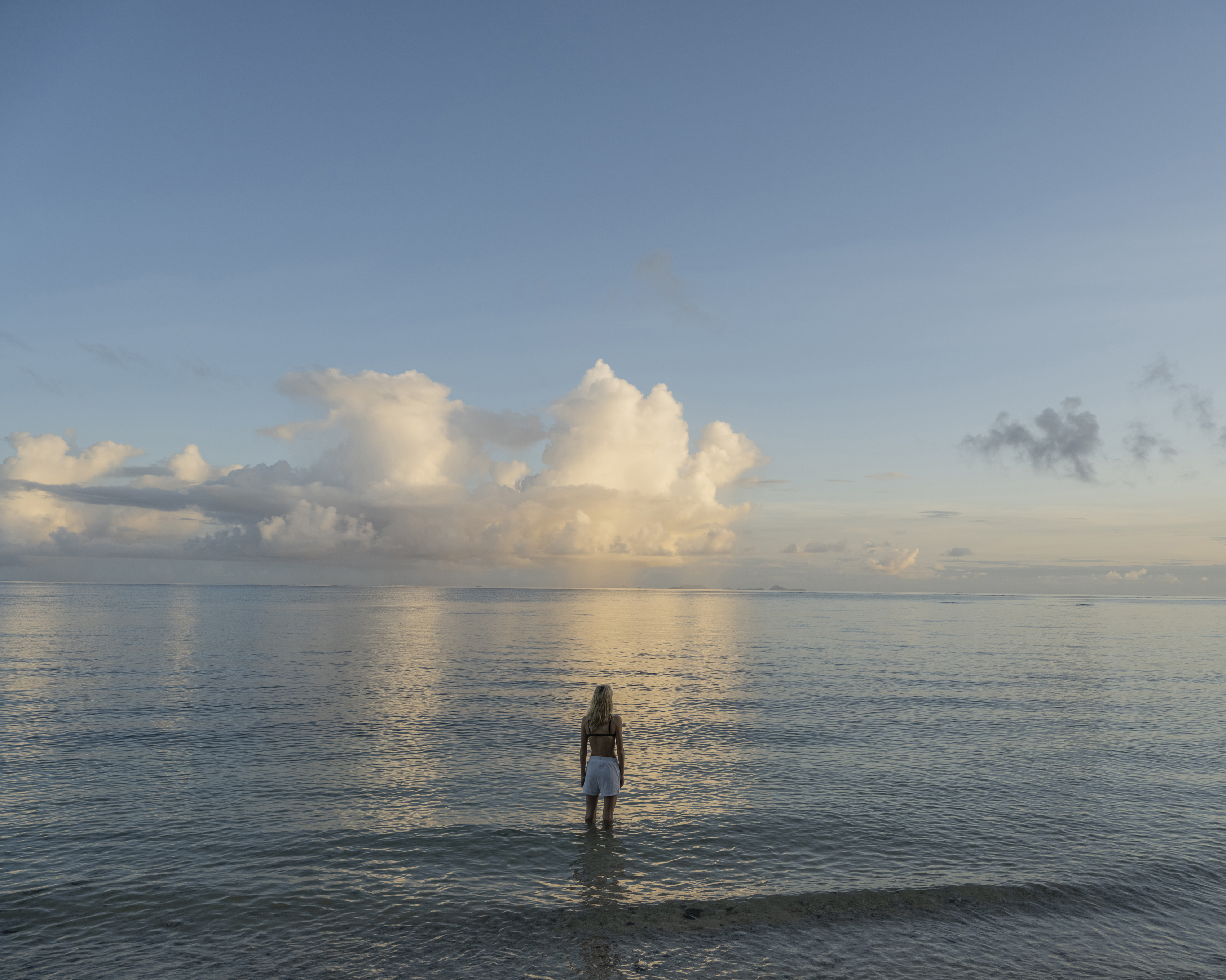 Woman stands in shallow ocean water at sunset. Clouds fill sky with blue hues. Long shot landscape composition. Tranquil scene.