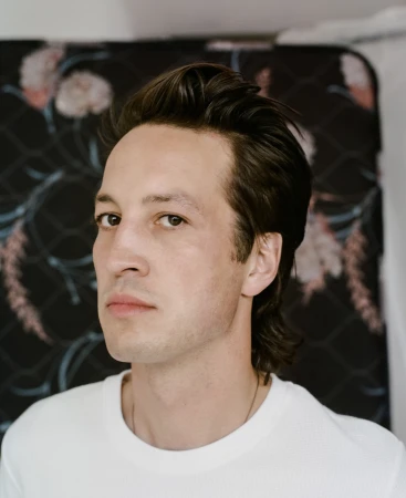 Close-up portrait of a dark-haired adult man with a neutral expression wearing a white t-shirt against a floral backdrop.