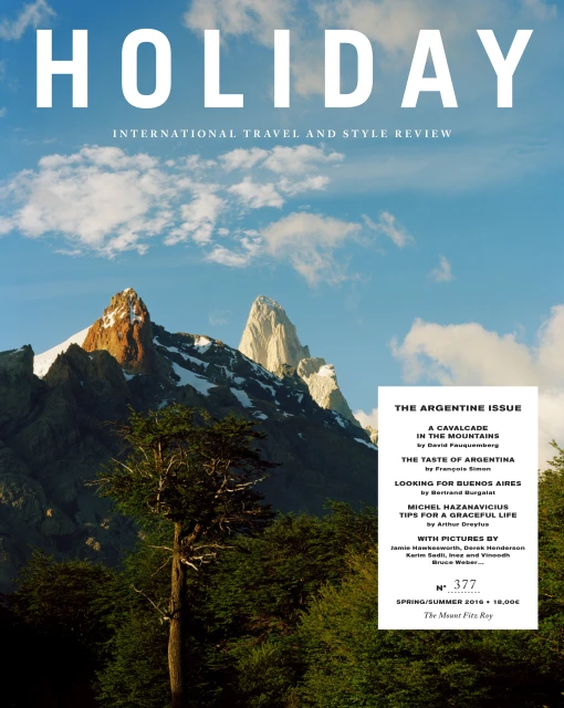 Holiday Magazine cover showcasing a majestic mountain range under a bright blue sky, surrounded by lush green forests. Travel and style publication.