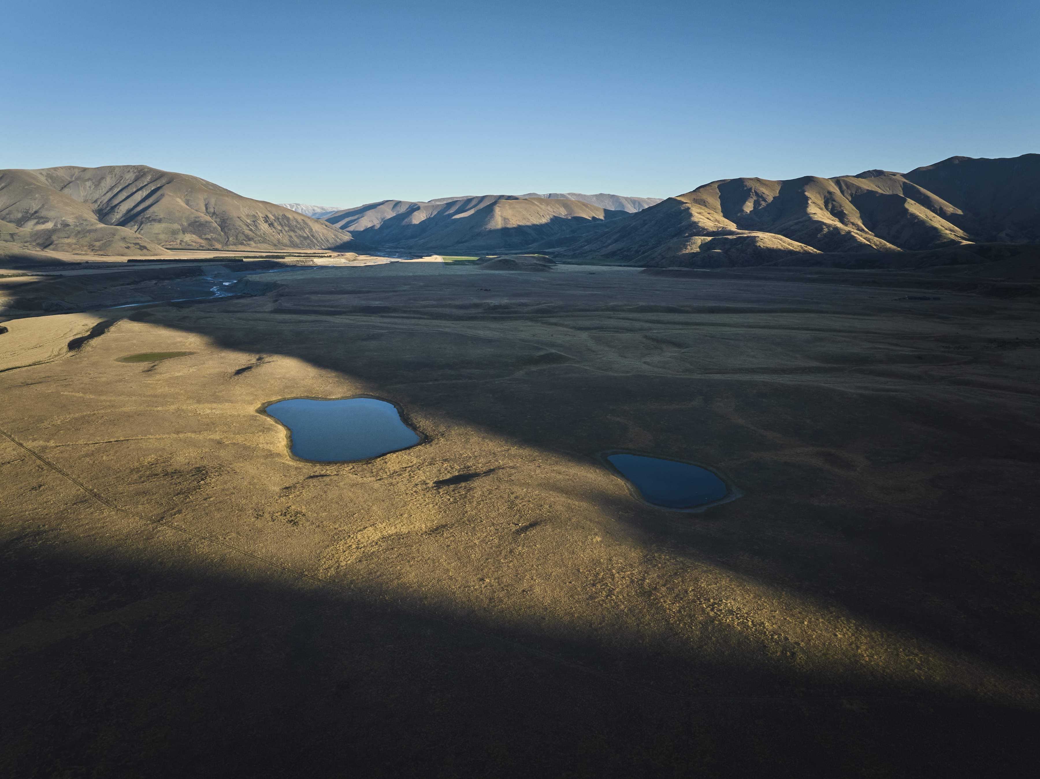 Aerial landscape photograph showing two small blue lakes in a vast dry valley surrounded by rolling mountains under a clear blue sky.