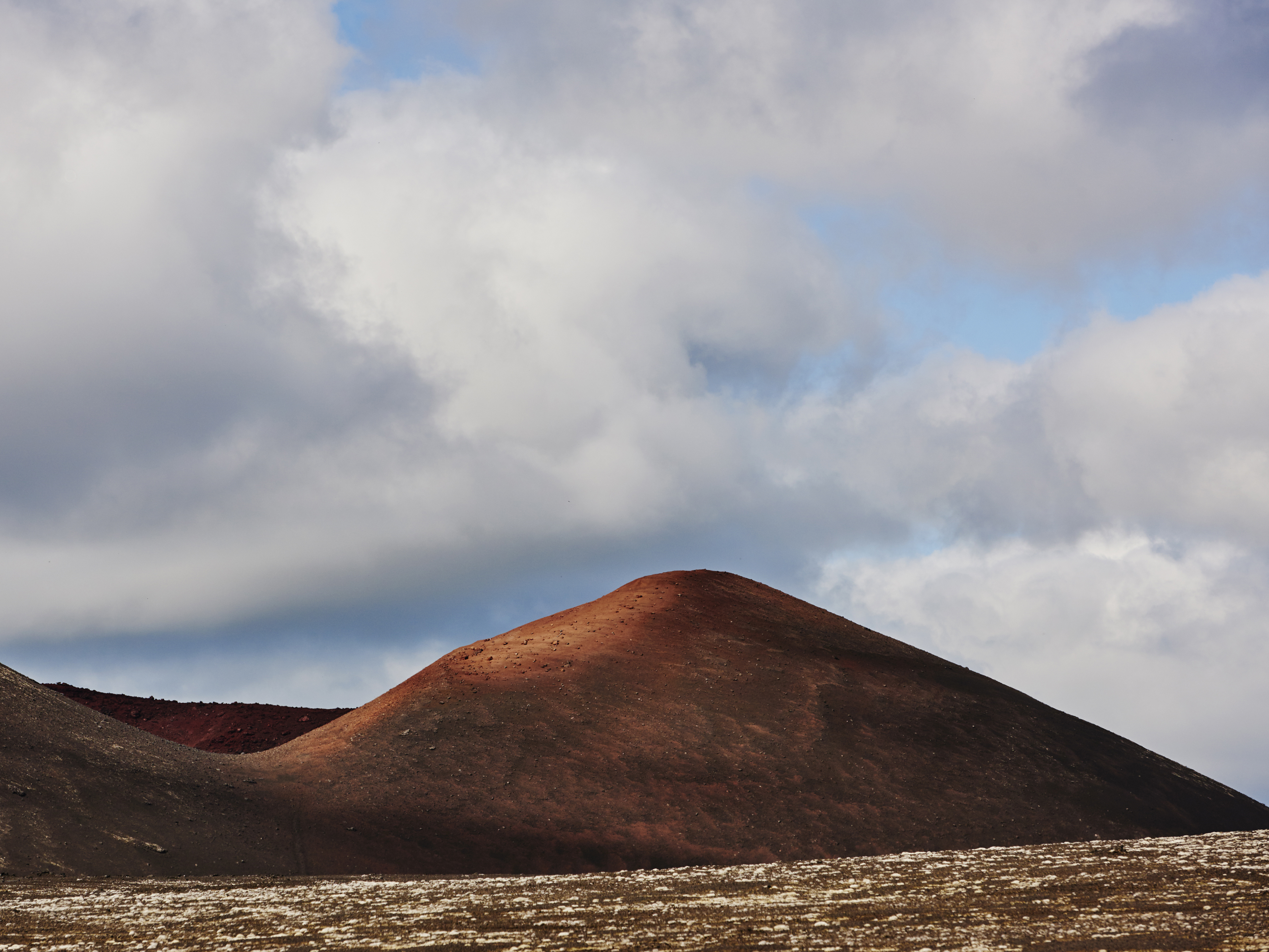 Landscape view of red and black volcanic hills under a cloudy sky, showcasing natural geological formations and unique terrain.