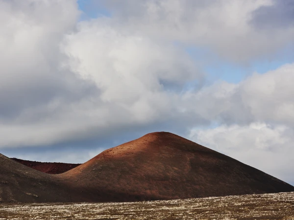 Landscape view of red and black volcanic hills under a cloudy sky, showcasing natural geological formations and unique terrain.
