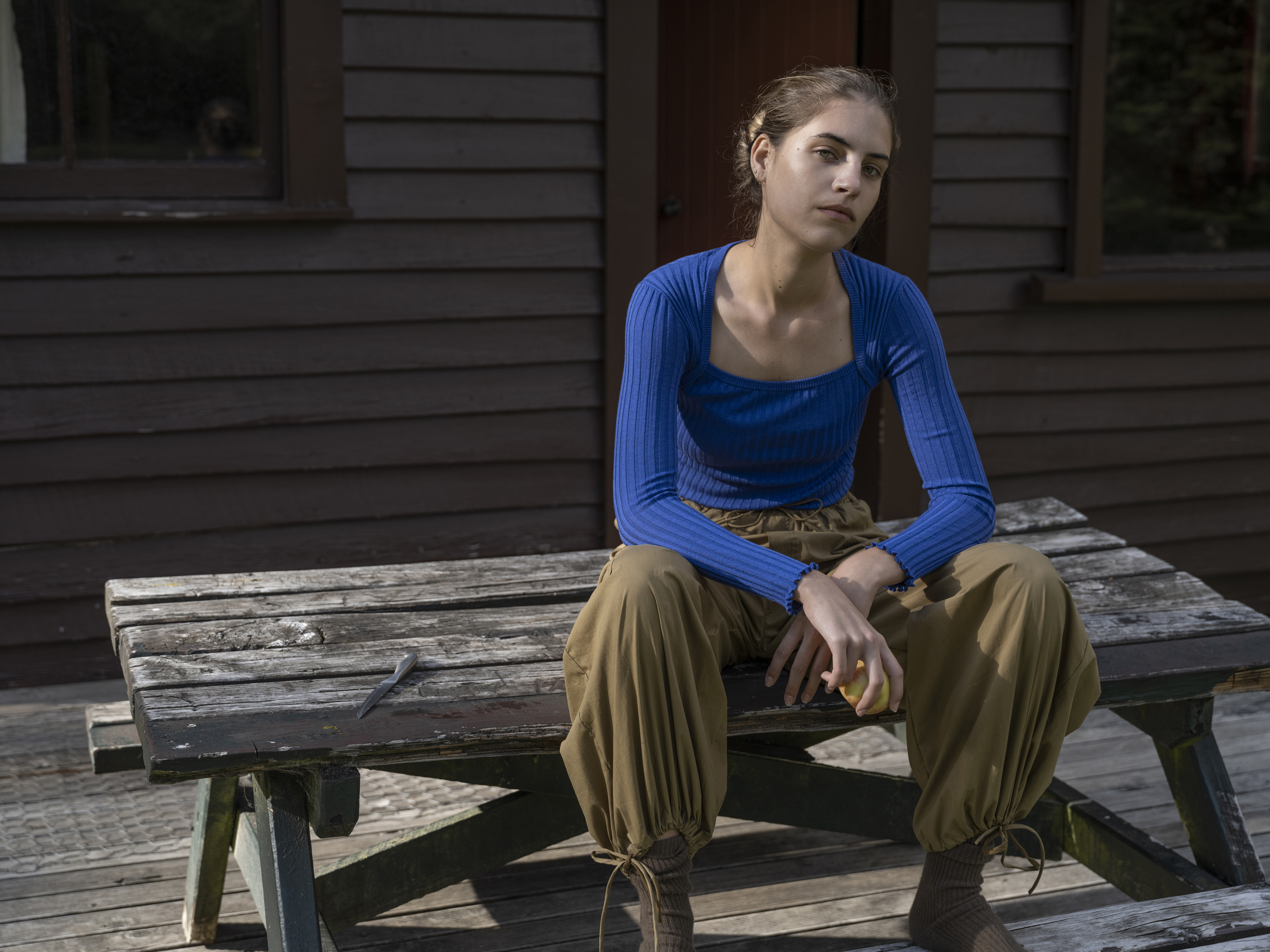 Young woman in a blue ribbed top sitting on a weathered wooden picnic table outside a dark building in Sydney