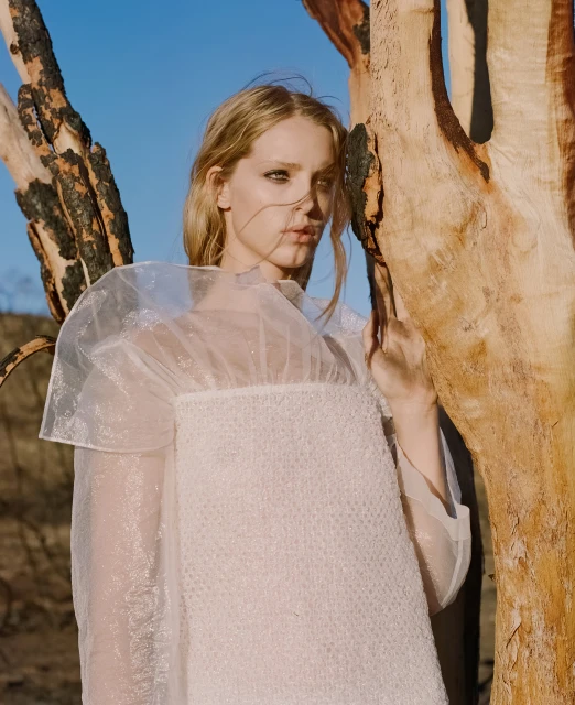 Blonde woman in white sheer dress poses next to a charred tree trunk against a clear blue sky in a fashion editorial shot.