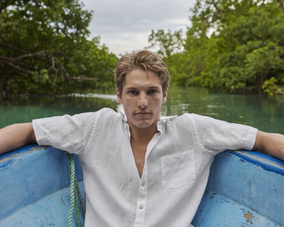 A young fair-skinned male in a white shirt sits in a blue boat traveling down a river with green foliage on either side beneath a cloudy sky.