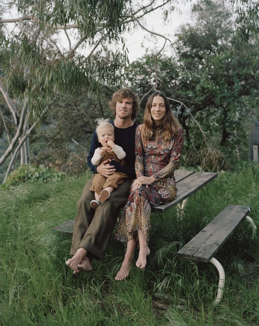 A family portrait shows a man, woman, and child sitting on a wooden park bench surrounded by tall grass in an outdoor setting.