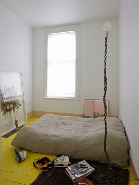 Minimalist interior with a simple mattress bed, chain floor lamp, and eclectic decor in a bright, white-walled room with a yellow floor.