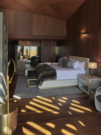 Luxurious modern bedroom interior with warm wood paneling, a large bed with fur throw, and patterned sunlight on wooden floor.