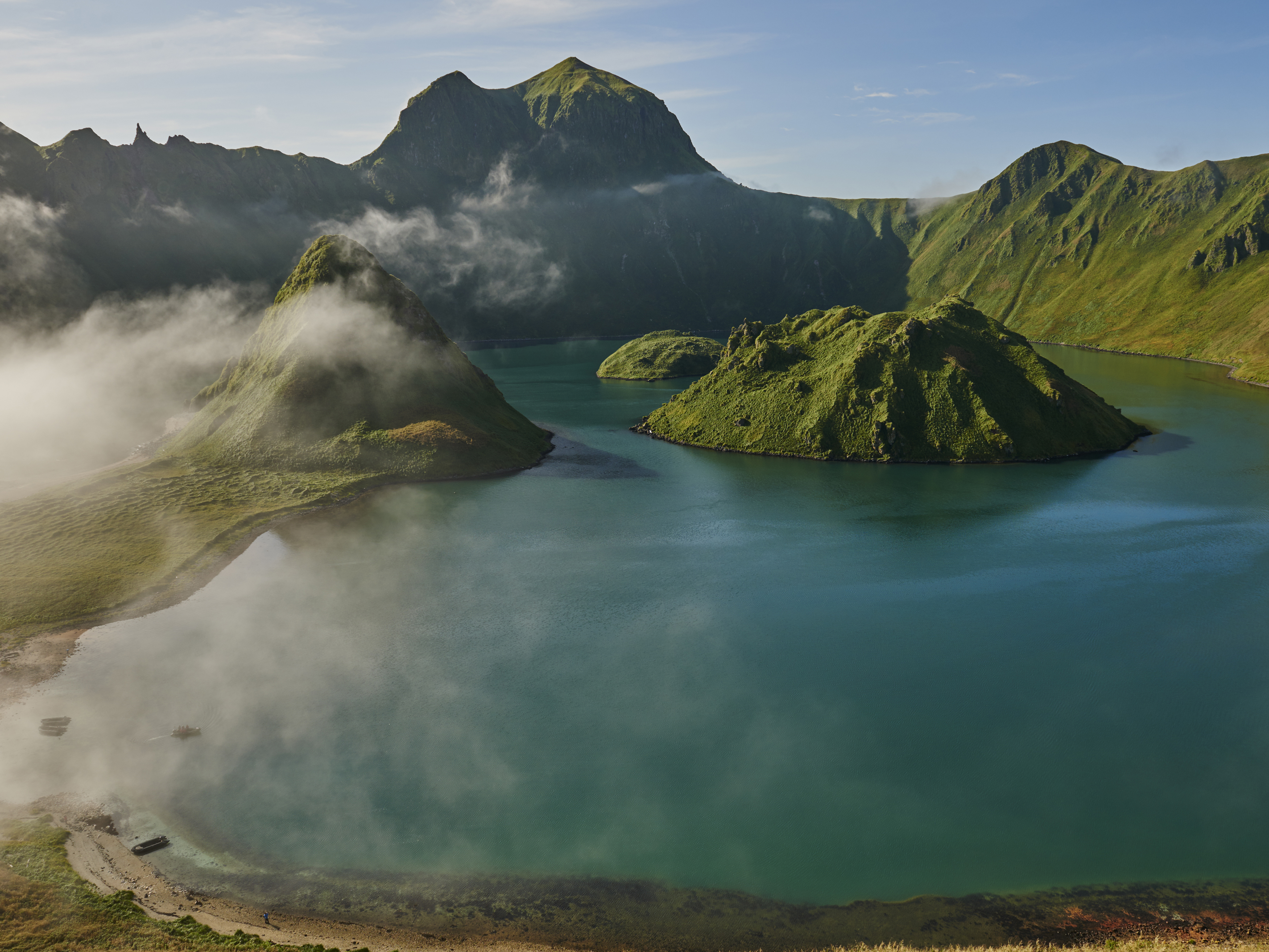 Overhead shot of turquoise lake with islands, surrounded by green mountains, some partially covered by fog on a sunny day.