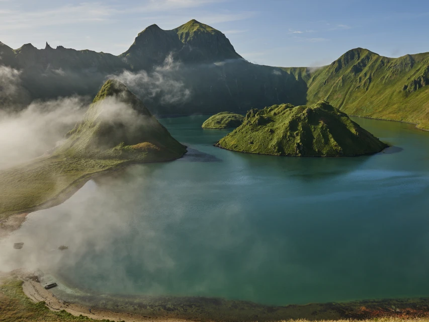 Overhead shot of turquoise lake with islands, surrounded by green mountains, some partially covered by fog on a sunny day.