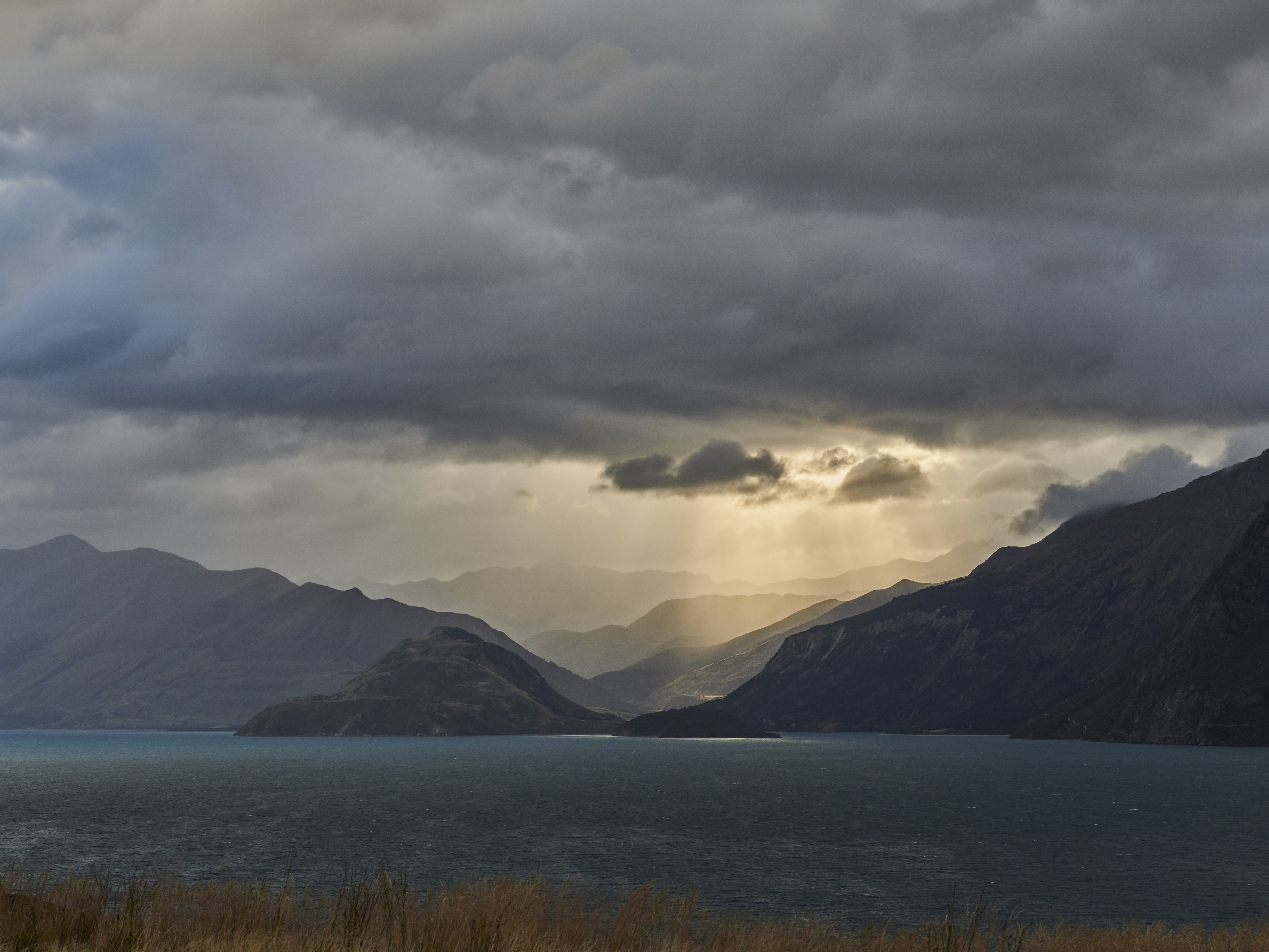 Scenic landscape featuring a tranquil lake surrounded by rolling hills and mountains under a cloudy sky with golden light shafts.