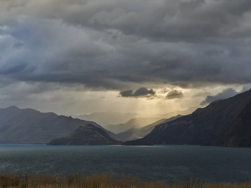 Scenic landscape featuring a tranquil lake surrounded by rolling hills and mountains under a cloudy sky with golden light shafts.