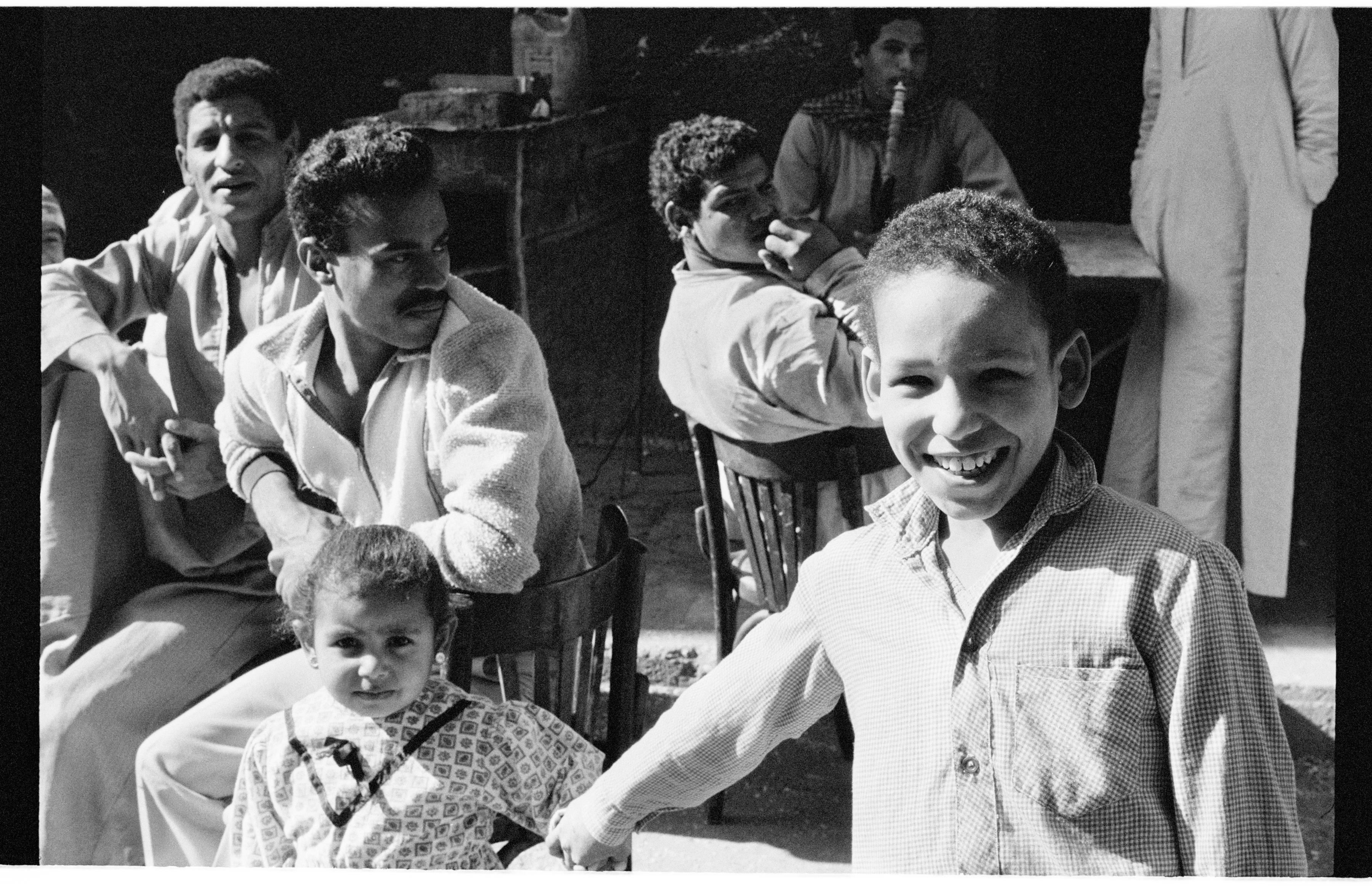 Black and white photo of a smiling young boy in a button-down shirt, with men and a young girl seated in the background.