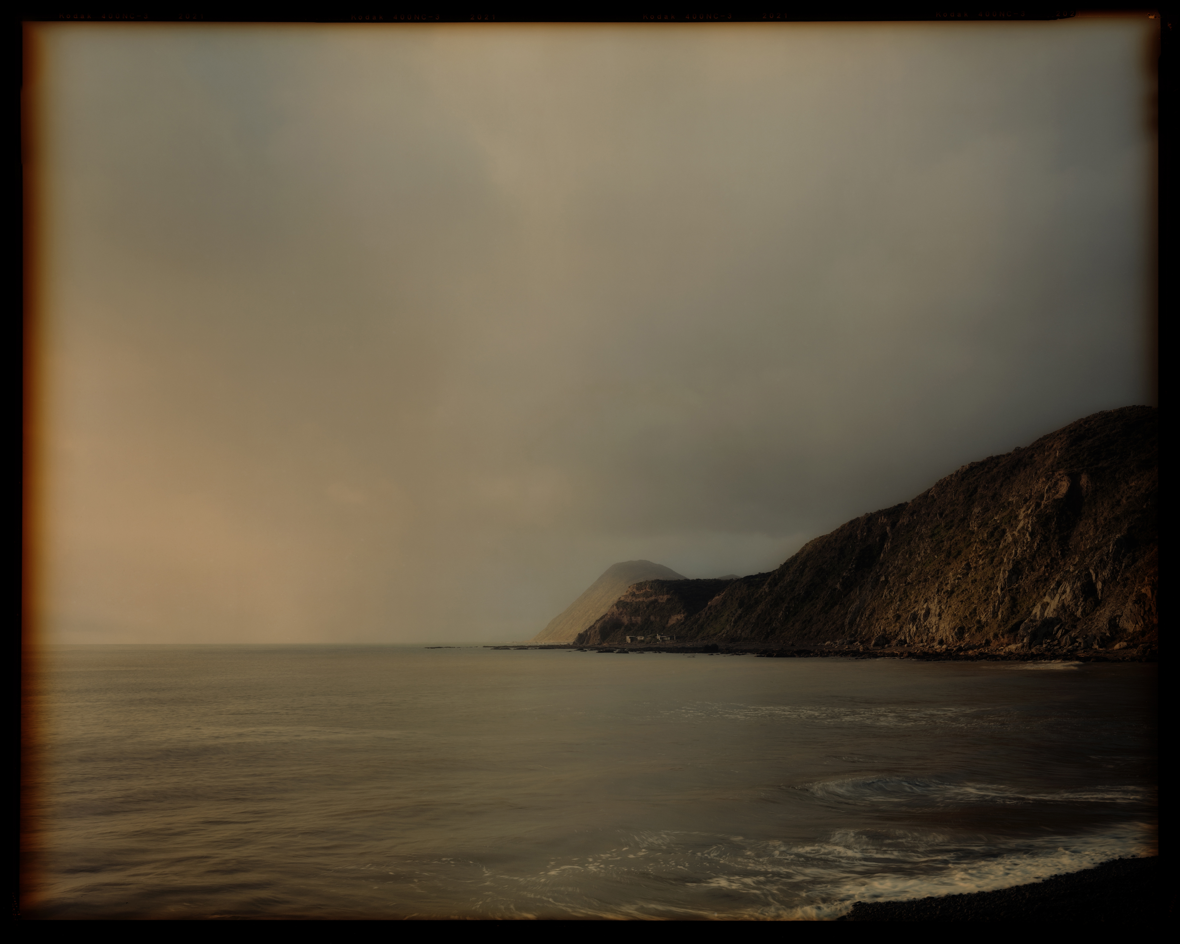 Coastal landscape featuring a tranquil sea, headland, and cloudy sky, evoking a sense of serene isolation in muted colors.