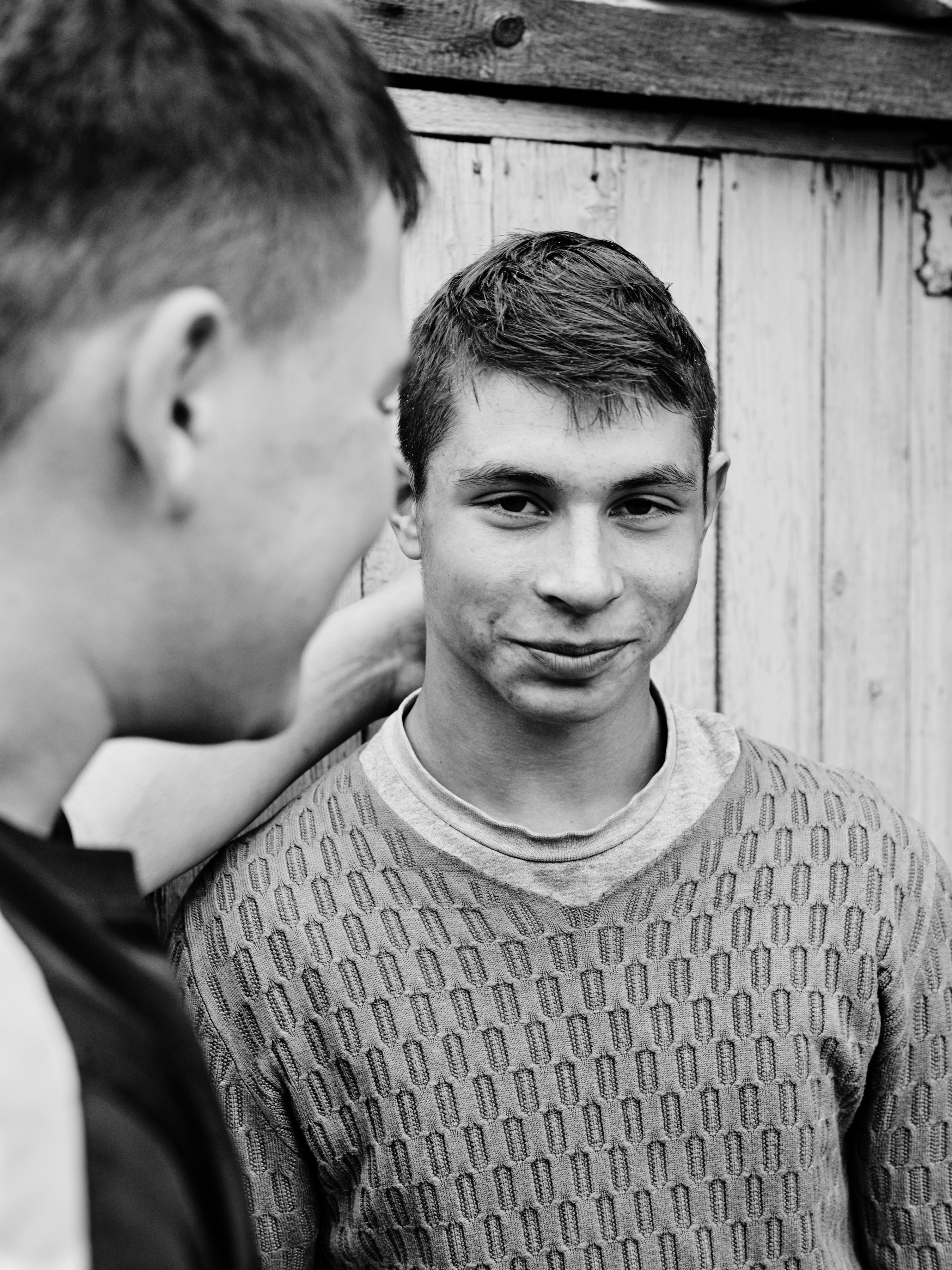 Black and white portrait of two young men outdoors, one with a patterned sweater, standing against a weathered wooden fence.