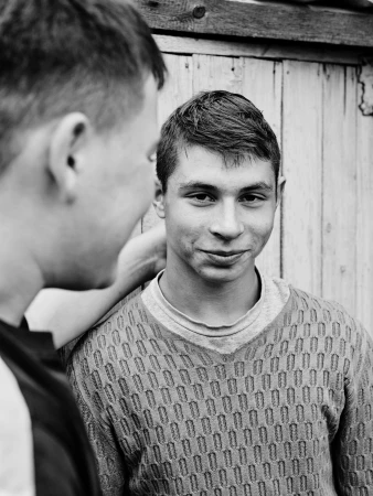 Black and white portrait of two young men outdoors, one with a patterned sweater, standing against a weathered wooden fence.