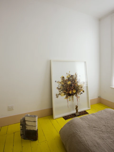 Bright interior photograph featuring a framed floral artwork leaning against a white wall, complemented by a bed and painted yellow floor.