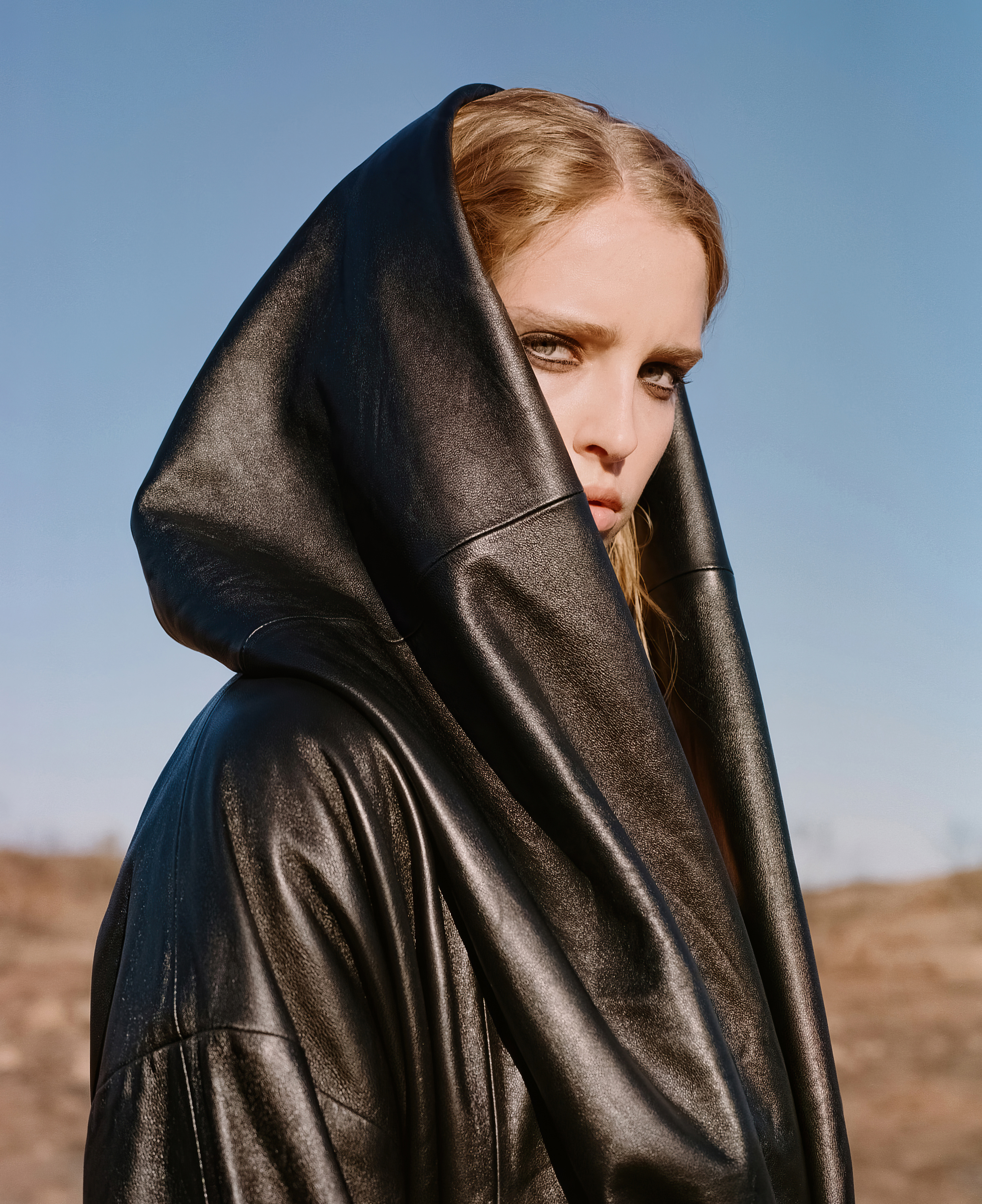 Woman in black leather hooded jacket gazes intensely at camera in an outdoor setting with a clear blue sky as a backdrop.