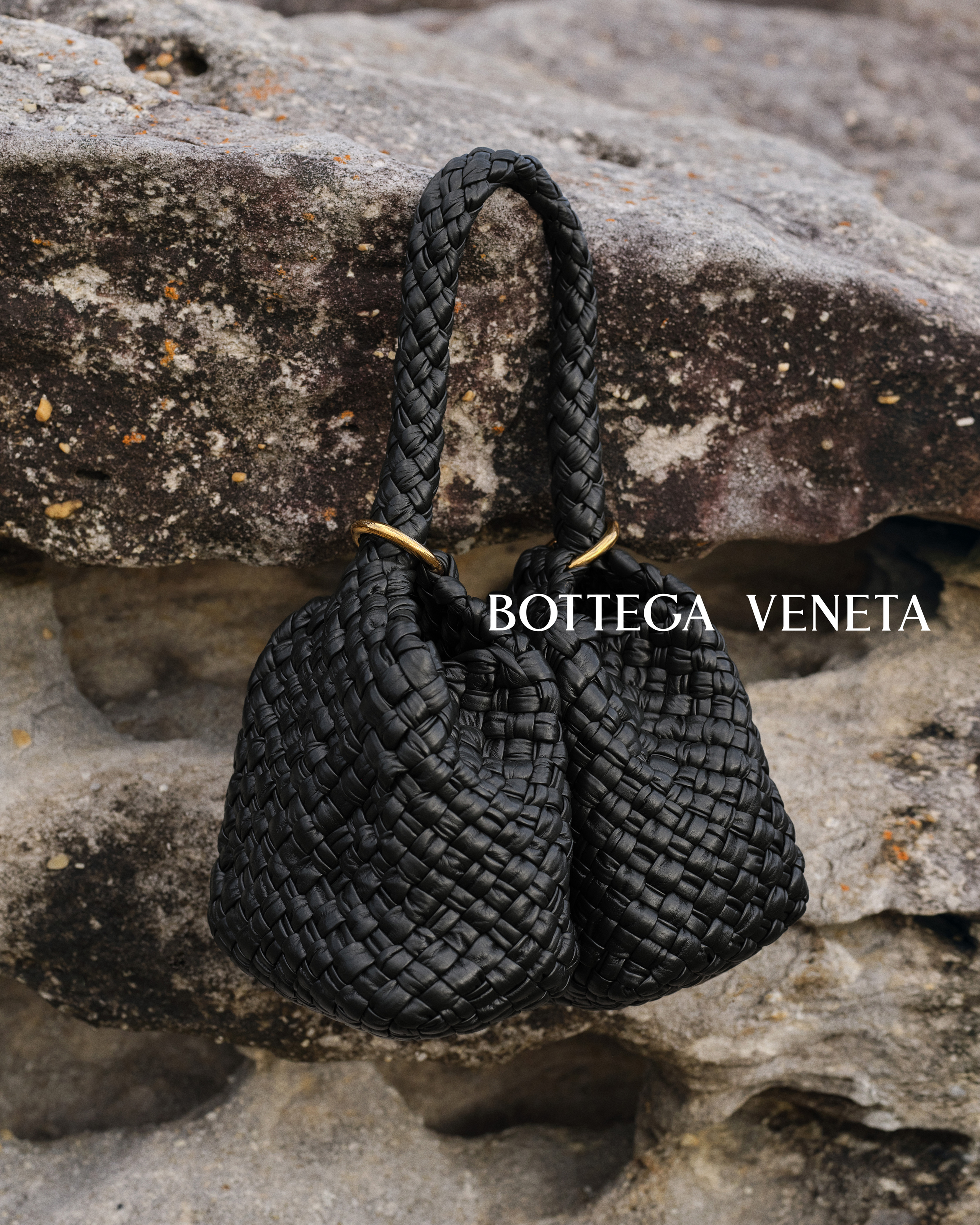 Woven black leather handbag from Bottega Veneta with braided top handle, against a rock background in an outdoor setting.