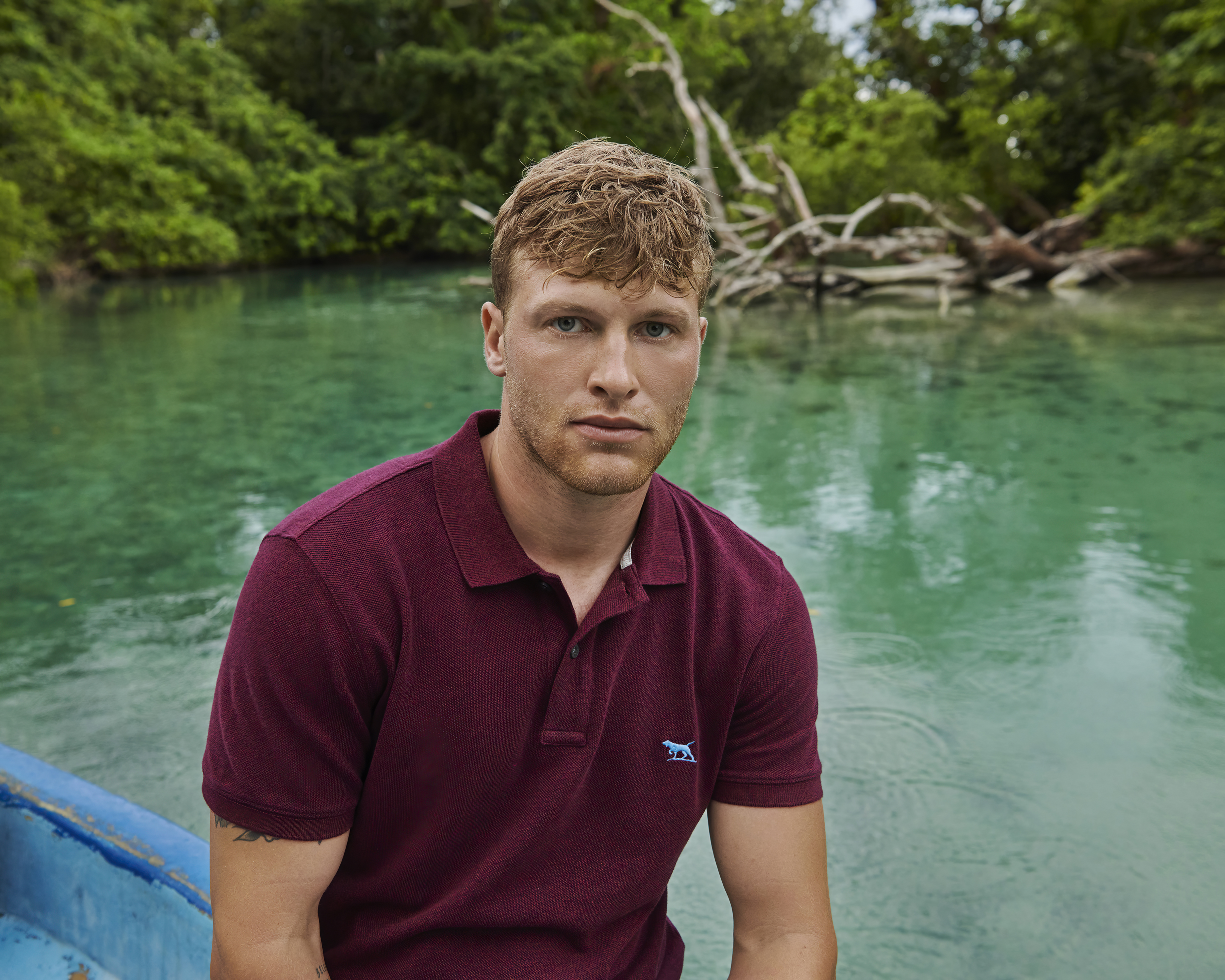 Young man with auburn hair wearing a maroon polo shirt seated in a blue boat, with a background of green water and lush trees.