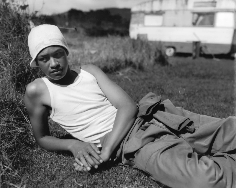 Portrait of an adult lying on a grassy field next to a trailer; wearing a white tank top and hat in a black-and-white landscape.