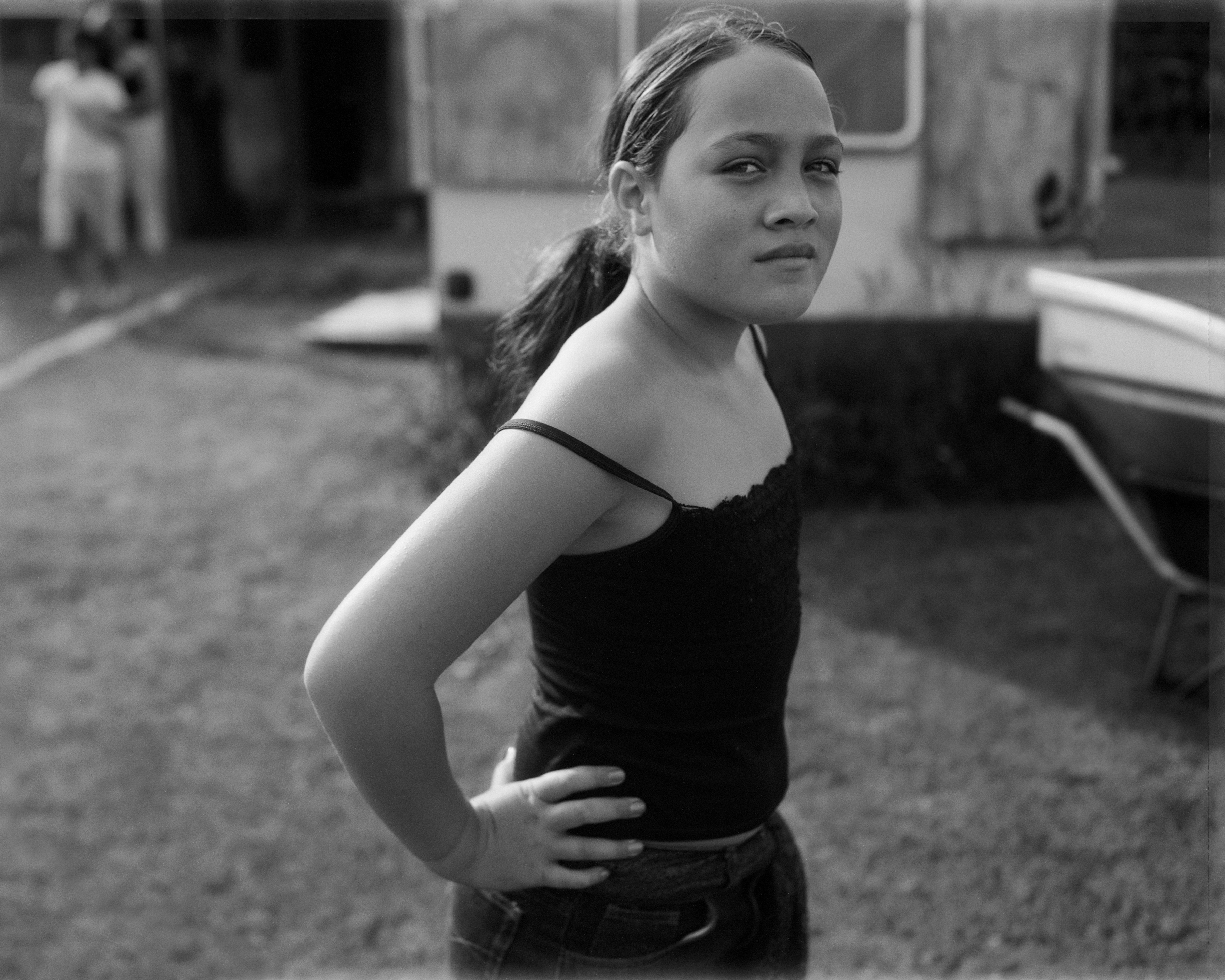 Monochrome portrait of a young person with a ponytail, standing outside with hands on hips near a trailer and a wheelbarrow.