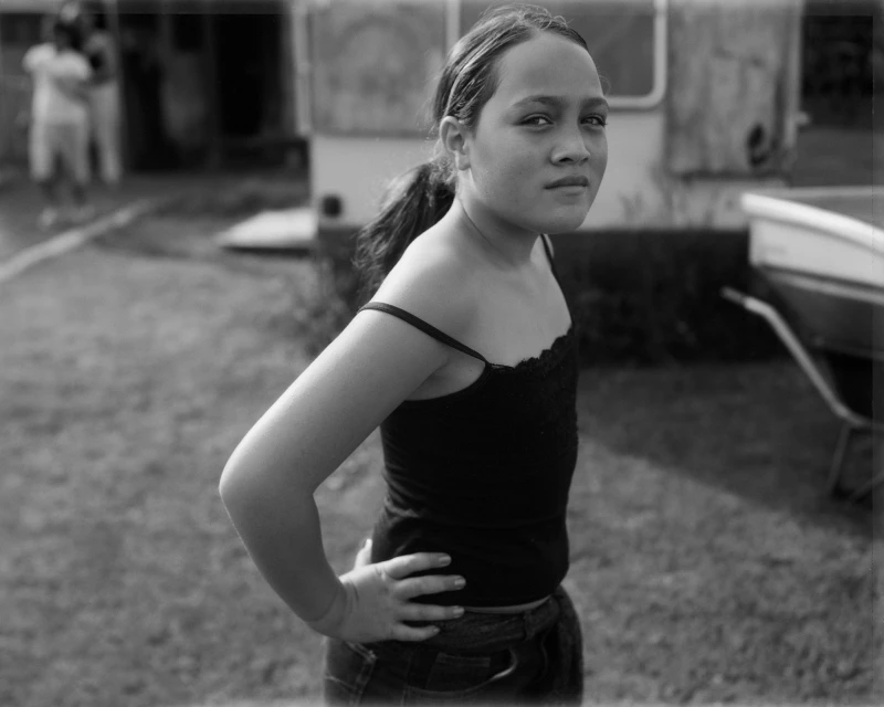 Monochrome portrait of a young person with a ponytail, standing outside with hands on hips near a trailer and a wheelbarrow.