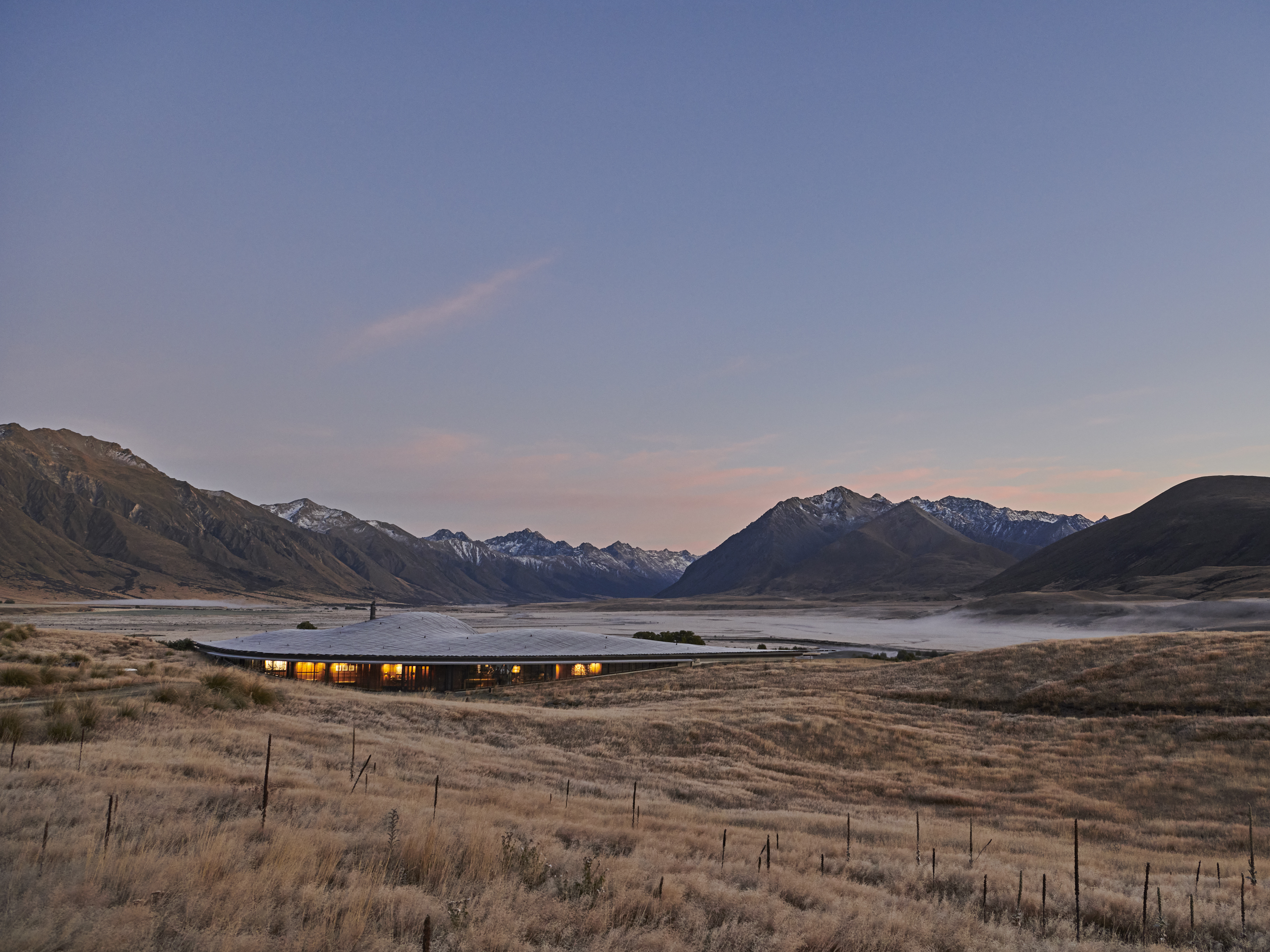 Exterior of a modern architectural building with a gently curved roof, set in a grassy field with snowy mountains in the background.