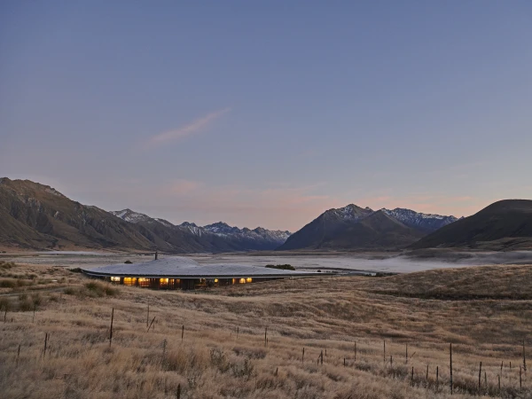Exterior of a modern architectural building with a gently curved roof, set in a grassy field with snowy mountains in the background.