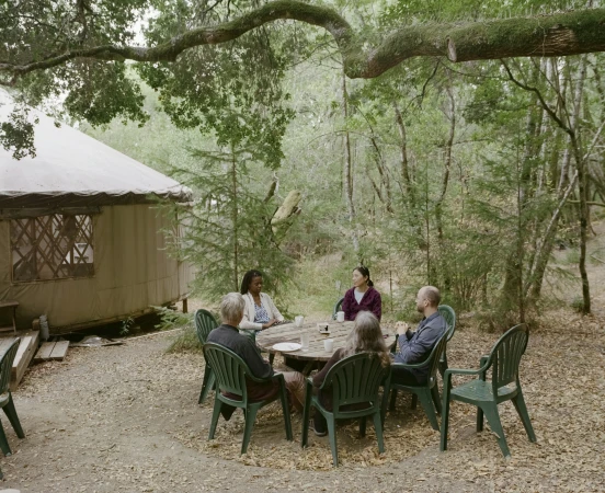 A group of five people sit around a wooden table outdoors in a wooded area with a tent in the background on a sunny day.