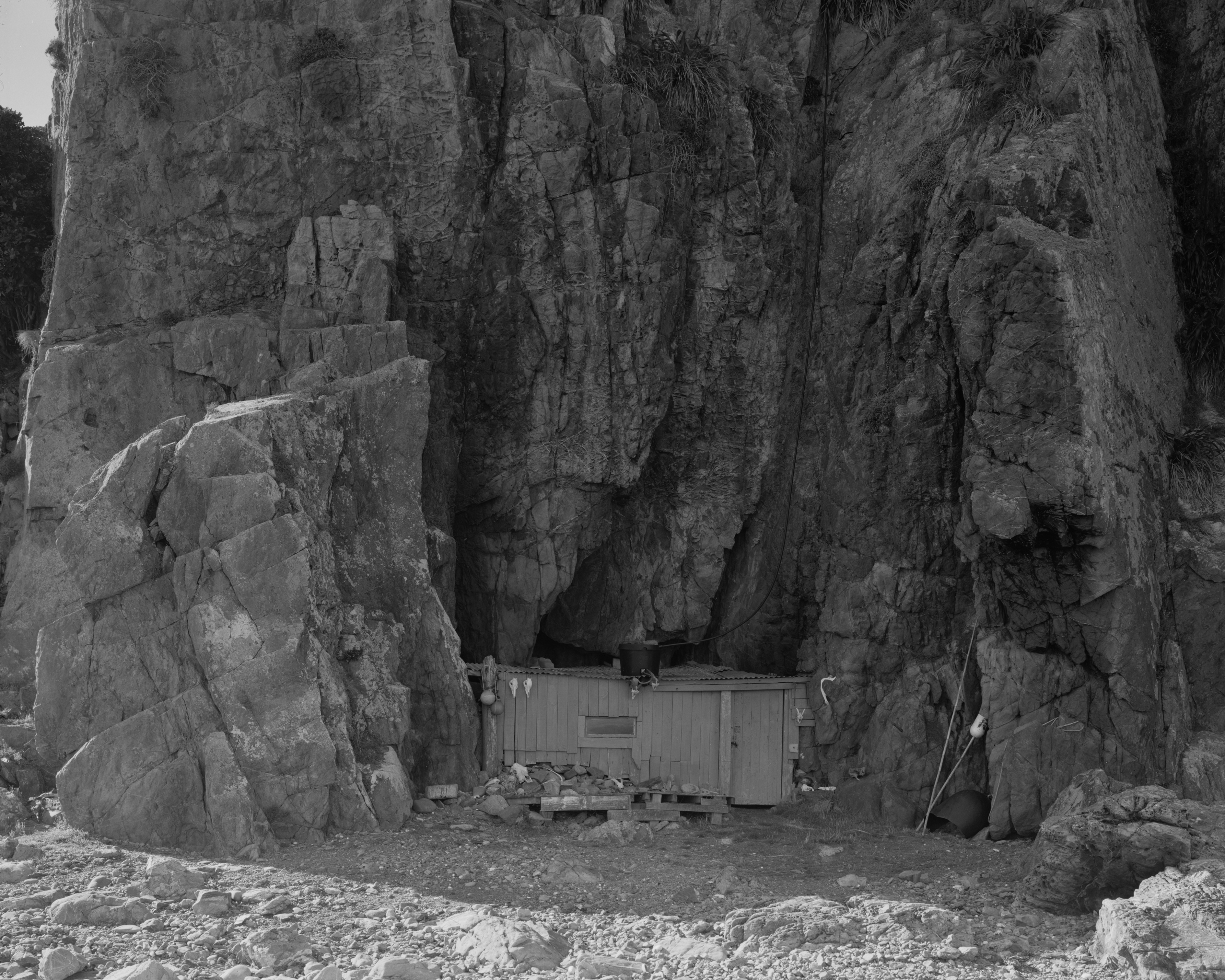 Monochrome landscape features a weathered wooden structure nestled at the base of a towering, rocky cliff, with scattered rocks in the foreground.