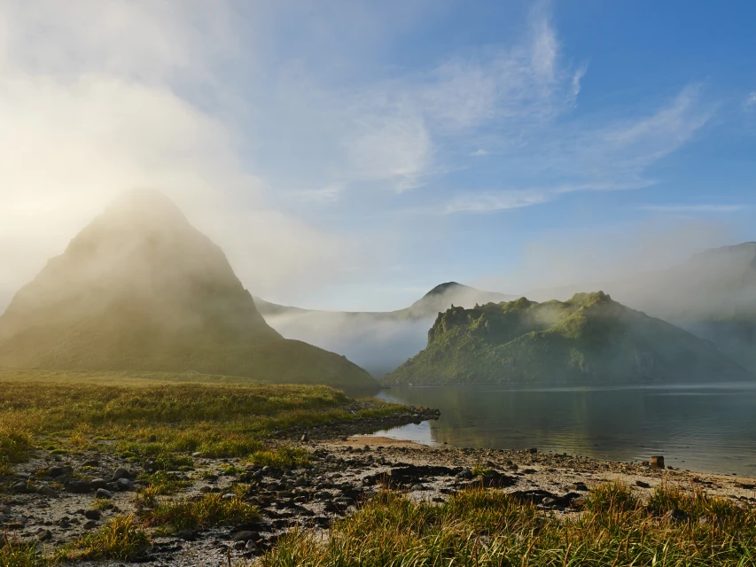 Scenic landscape featuring mountains partially obscured by low-lying fog, a calm body of water, and lush green vegetation.