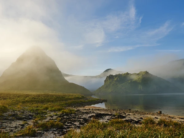 Scenic landscape featuring mountains partially obscured by low-lying fog, a calm body of water, and lush green vegetation.