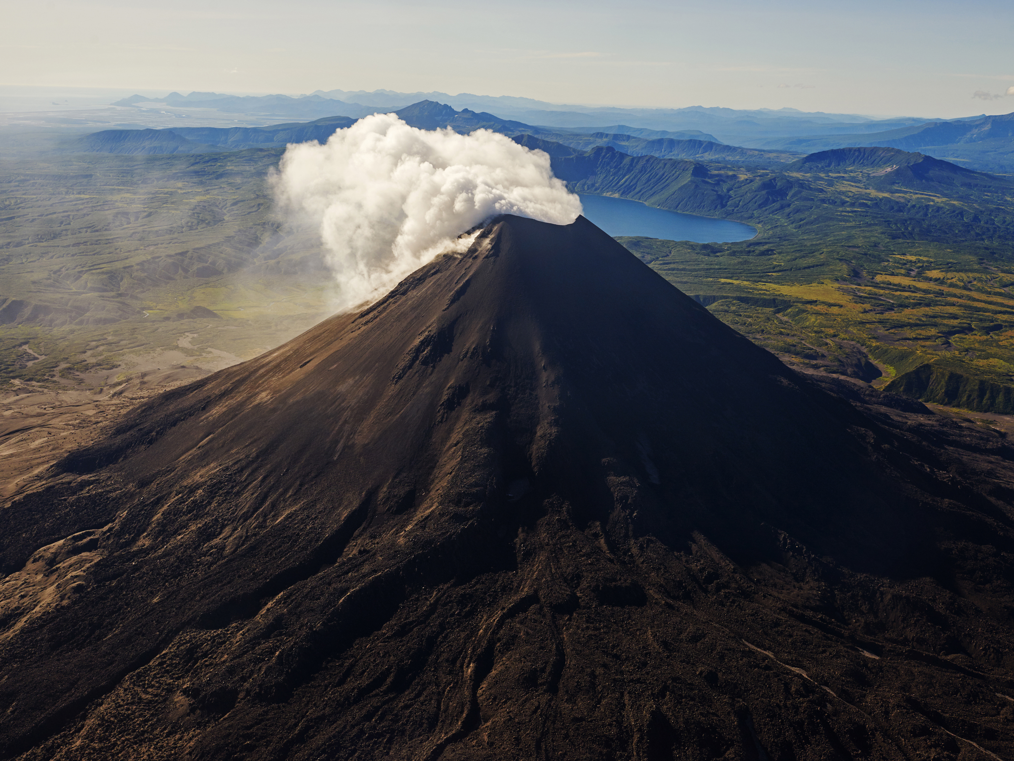 Aerial view of a striking dark volcano with a plume of white smoke rising from its peak, surrounded by a verdant landscape of mountains.