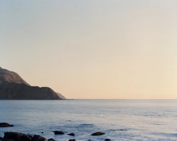 Coastal landscape with calm ocean waters, dark rocky shorelines, and distant rolling hills under a soft pastel sunset sky.