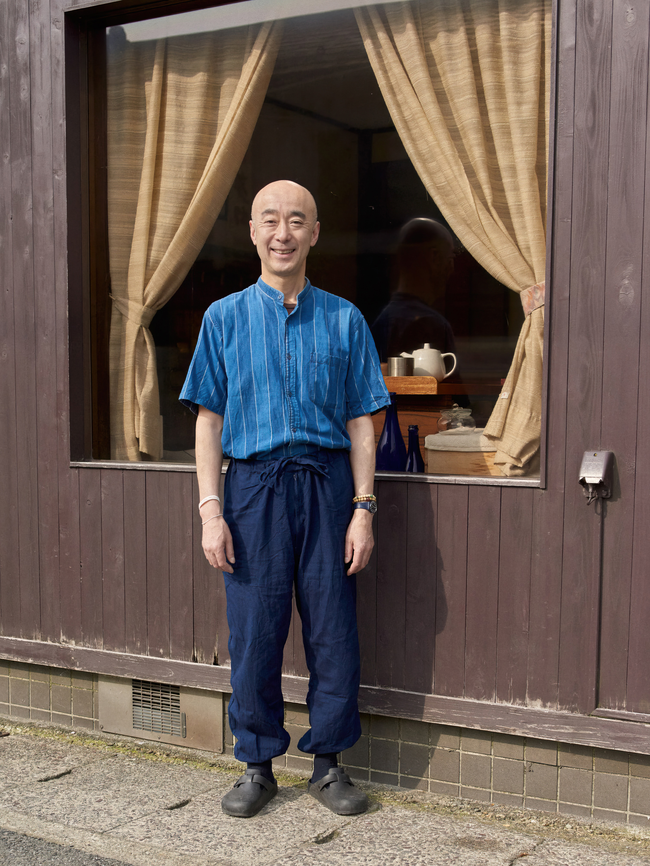 Bald adult man in blue shirt stands outside a wooden building with a window and tan curtains, facing camera with a slight smile.