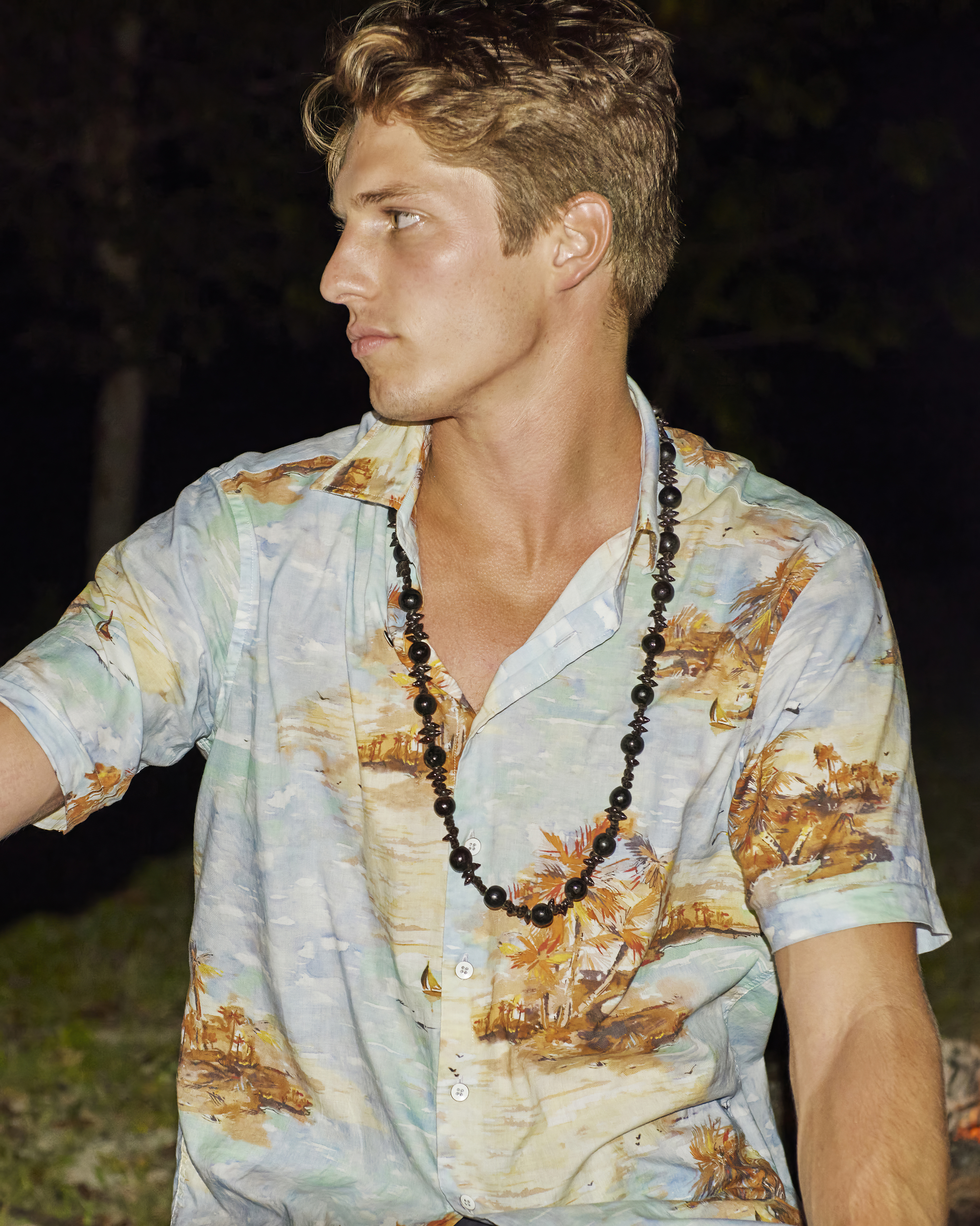 Portrait of a blond adult male outdoors, wearing a patterned short-sleeve shirt and beaded necklace, gazing off-camera into the distance.