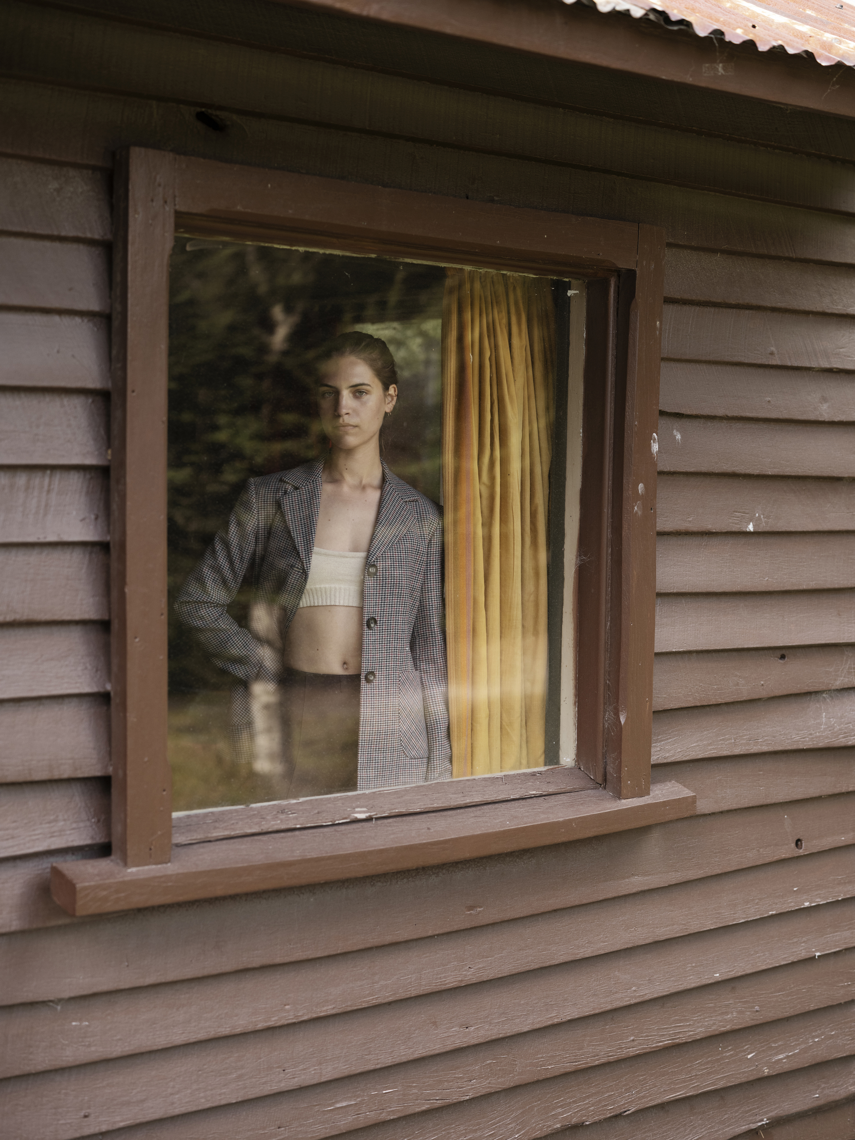Woman in grey plaid blazer and white crop top viewed through a wood cabin window by photographer Derek Henderson.