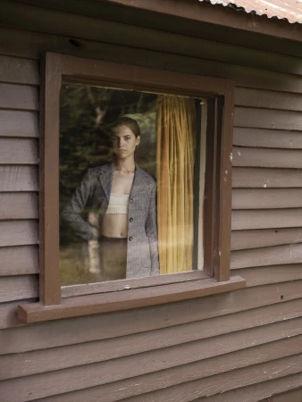 Woman in grey plaid blazer and white crop top viewed through a wood cabin window by photographer Derek Henderson.