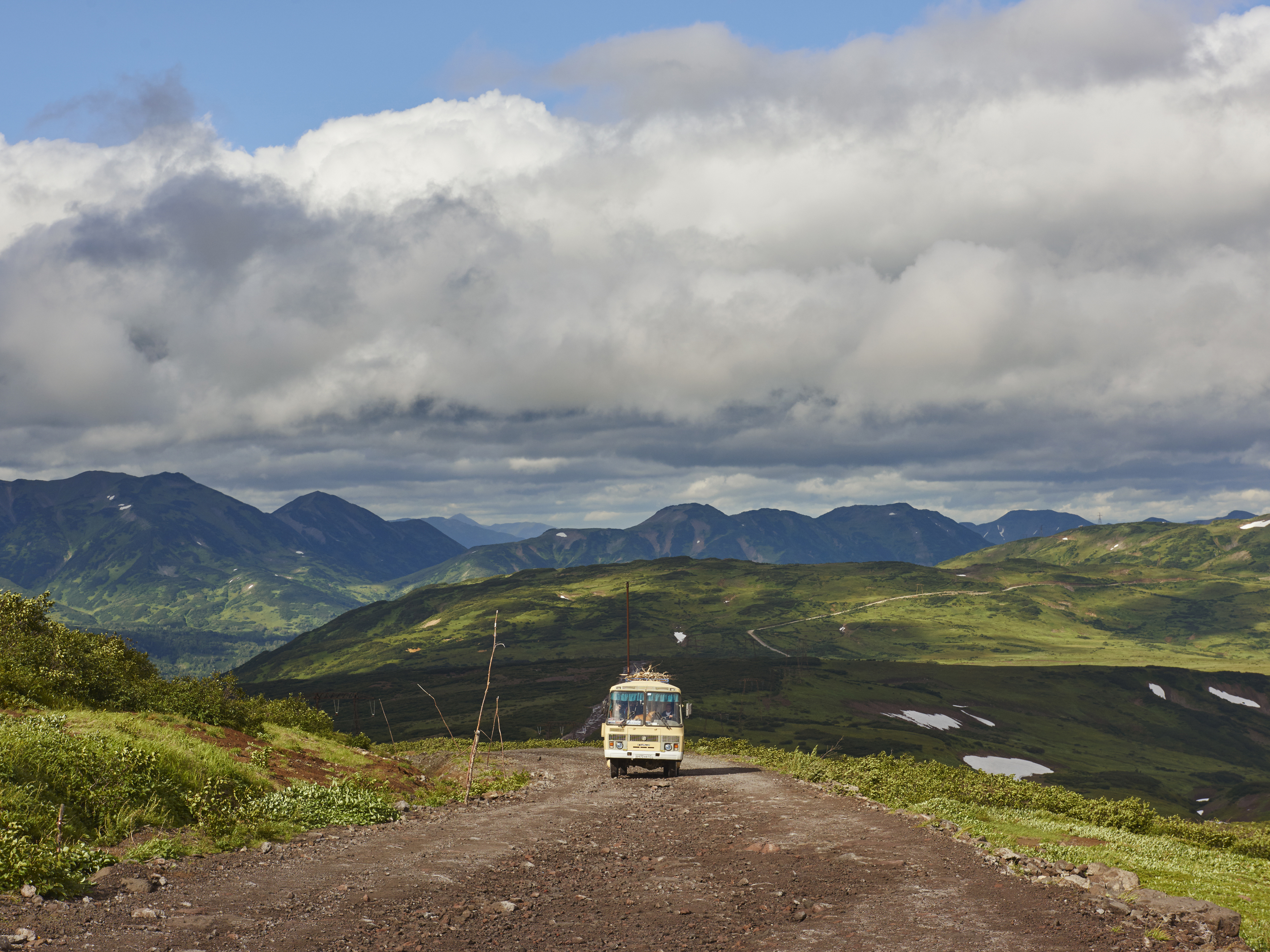 A vintage bus travels on a dirt road through a hilly landscape with cloudy skies, showcasing travel and adventure in nature.