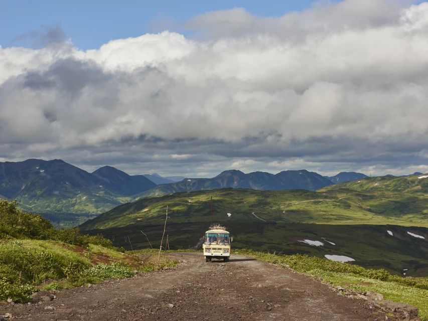 A vintage bus travels on a dirt road through a hilly landscape with cloudy skies, showcasing travel and adventure in nature.